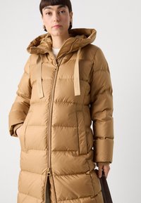 Langer Puffer-Mantel in einem warmen Beige, mit gepolstertem Textur, Kapuze und Frontreißverschluss. Verfügt über verstellbare Träger und Seitentaschen.