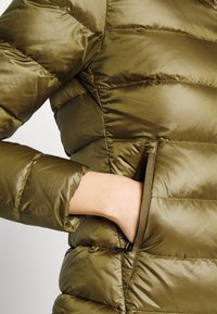 Chaqueta acolchada de color verde oliva con una textura lisa y brillante. Cuenta con costuras horizontales y bolsillos laterales, con un puño ajustado en la manga.