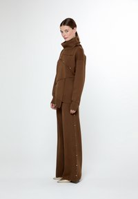 Kvinna i brun höghalssweater och vida byxor med guldknappdetaljer, iförd metallic spetsiga skor, stående mot en enkel vit bakgrund.