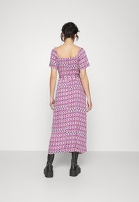 Robe maxi en violet avec un motif géométrique en diamants, manches courtes évasées, taille cintrée et texture douce. Associée à des bottes noires imposantes.