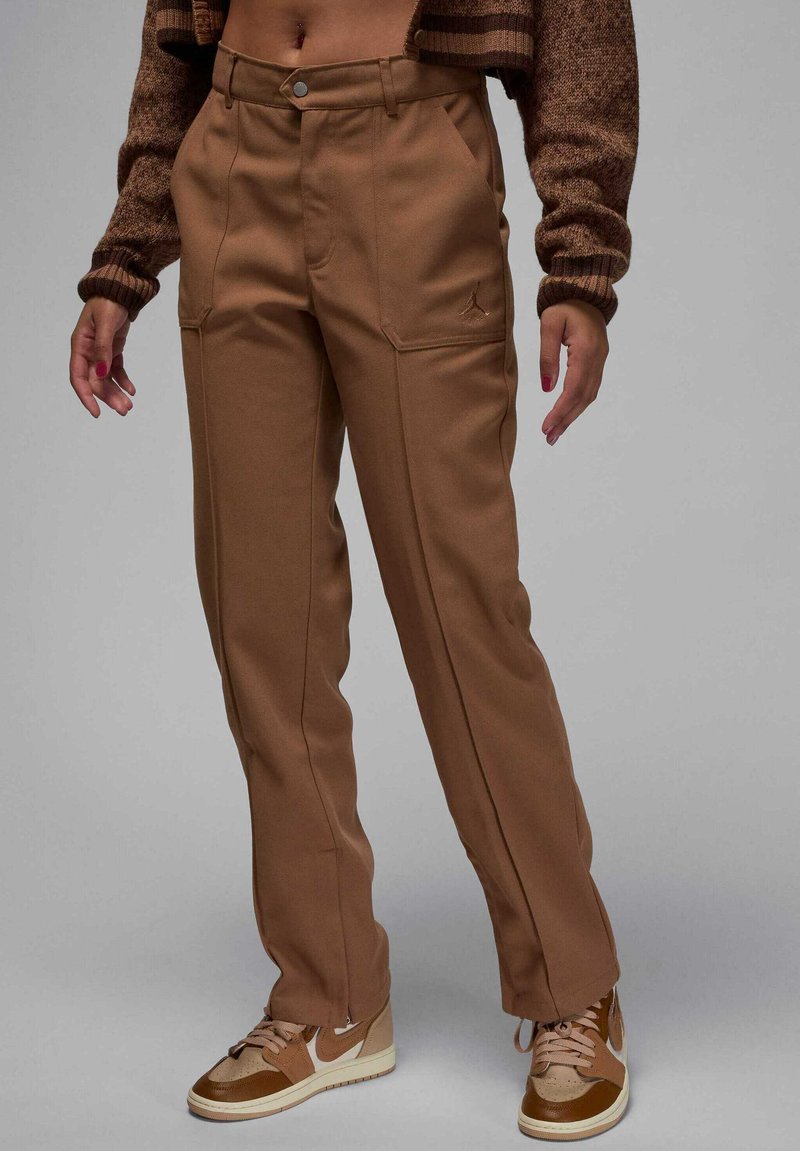 Persona che indossa pantaloni marroni a vita alta con tasche anteriori, un maglione corto marrone e sneaker Nike marroni e beige su uno sfondo grigio.