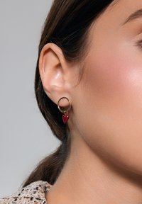 Złoty kolczyk w kształcie koła z małym, czerwonym serduszkiem, prezentujący błyszczące wykończenie i gładką fakturę. Subtelny i lekki design.