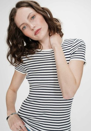 Femme aux cheveux bruns ondulés portant un t-shirt à manches courtes blanc à rayures noires, tenant sa nuque d'une main, sur un fond uni.