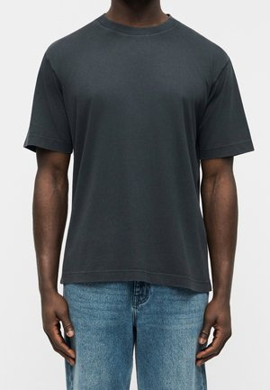 Homme portant un t-shirt gris foncé uni à manches courtes et un jean bleu clair, debout devant un fond clair uni.