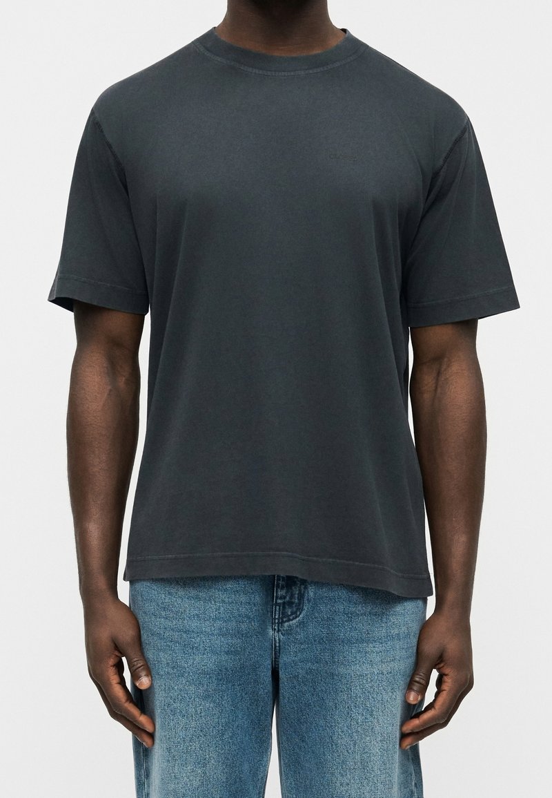 Homme portant un t-shirt gris foncé uni à manches courtes et un jean bleu clair, debout devant un fond clair uni.