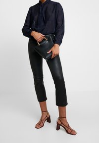 Osoba v průhledné námořnické blůze, černých zkrácených kožených kalhotách, hnědých sandálech na podpatku, držící černou kabelku se zlatými úchyty.