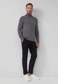 Man staat tegen een muur gekleed in een grijze coltrui, zwarte broek en witte sneakers, met zijn handen in zijn zakken.