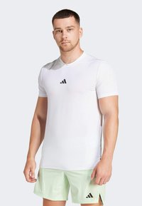 White athletic t-shirt made of breathable fabric, featuring short sleeves and a black logo on the chest, paired with light green shorts.