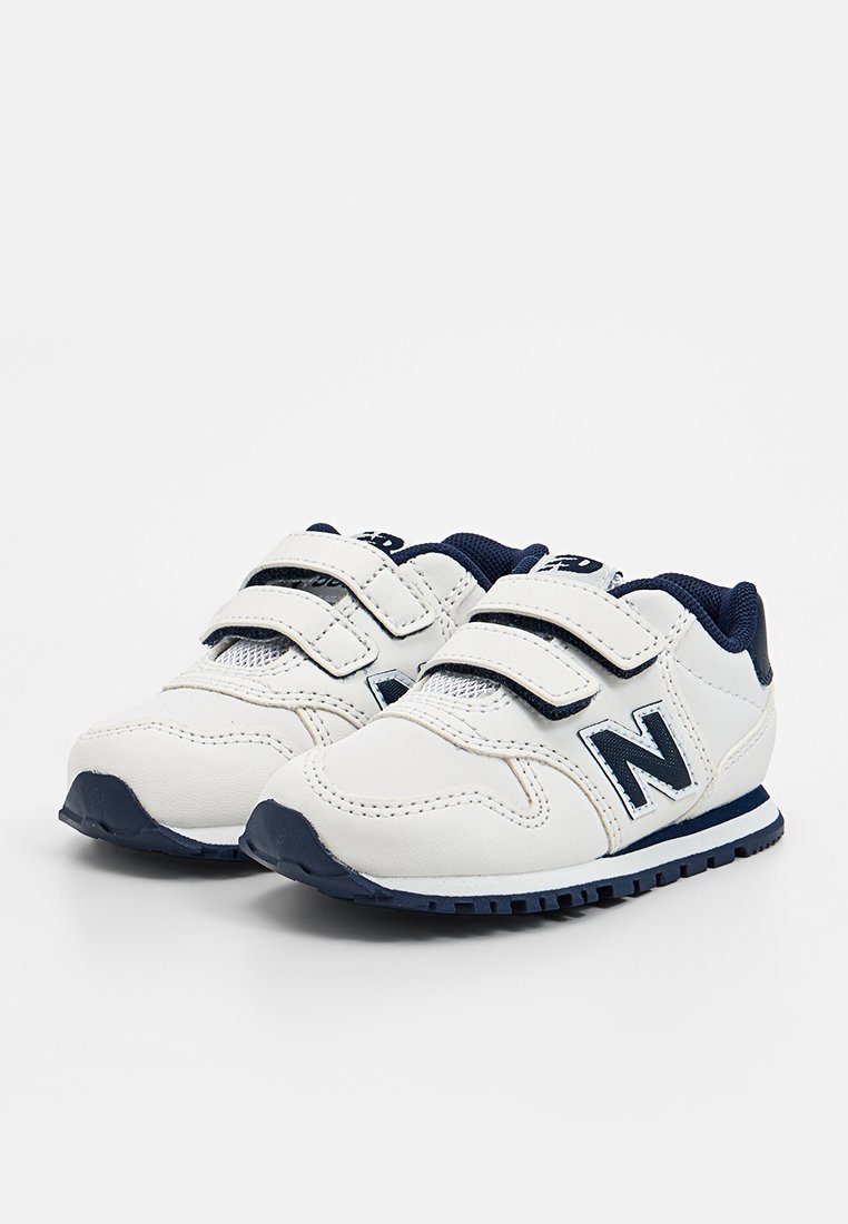 Baskets blanches à scratch avec des accents navy, présentant un dessus texturé, des panneaux en mesh et une semelle foncée contrastante. Le logo New Balance est visible.