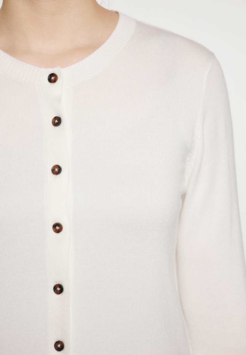 Witte gebreide cardigan met ronde hals, voorzien van een rij donkerbruine, ronde knopen aan de voorkant en geribbelde manchetten.