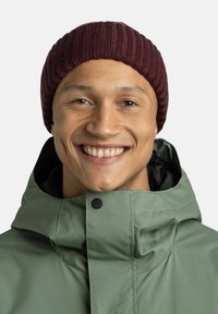 Maroon ribbed knit beanie atop a smiling individual wearing a green waterproof jacket with a high collar and black interior.