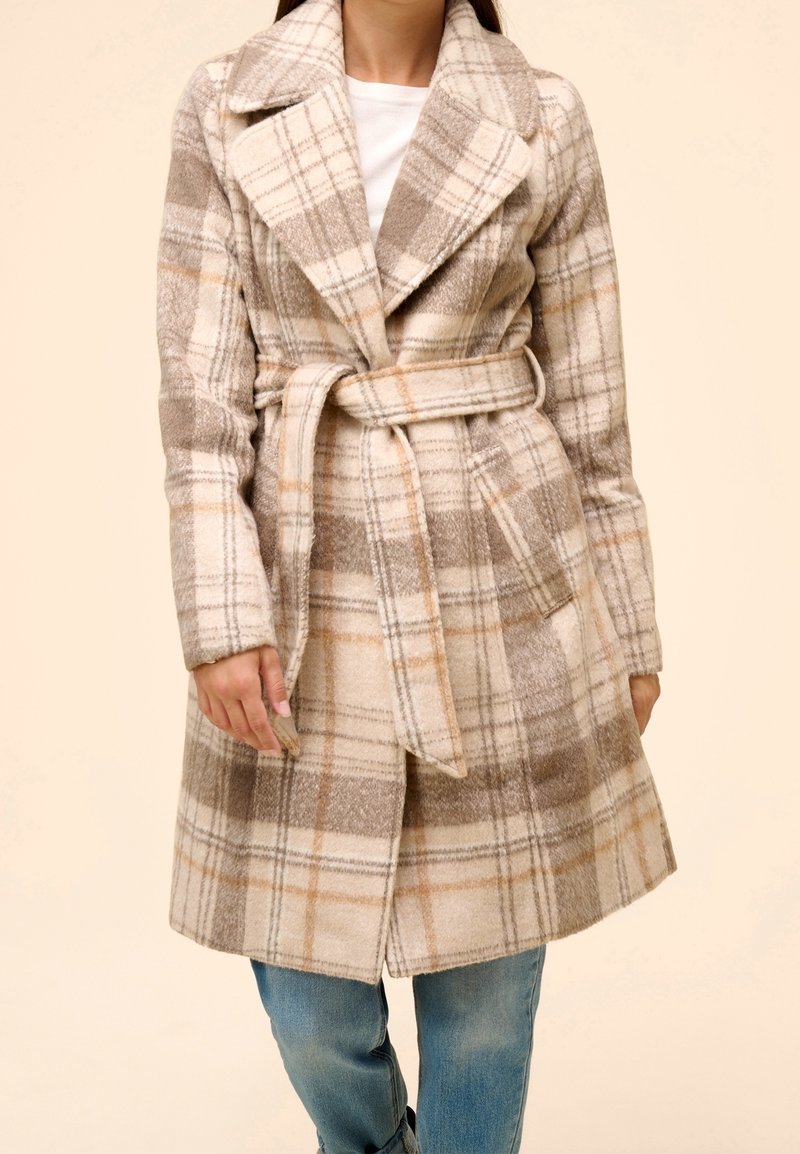 Femme portant un manteau en laine beige et marron à carreaux ceinturé, par-dessus une chemise blanche et un jean bleu, debout sur un fond clair uni.