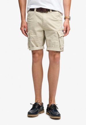 Man draagt beige cargoshorts met opgerolde zoom, wit overhemd, bruine riem, horloge en zwarte bootschoenen met lichtbruine zolen.
