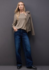 Maglione tan, trench coat grigio chiaro, jeans a gamba larga blu scuro e scarpe nere, posato su uno sfondo grigio. Texture lisce, vestibilità casual.