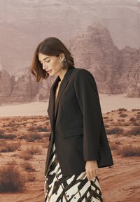 Juodas pritaikytas švarkas su išpjautomis apykaklėmis, vienu mygtuku ir kišenėmis, derinamas su raštuotu juodai baltu sijonu, dėvimas ant dykumos fono.