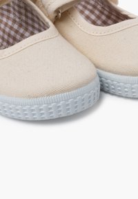 Scarpe di tela color beige chiaro con punta arrotondata, suola in gomma bianca e fodera a quadretti. Presentano una fascia con una fibbia metallica.