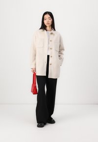 Fleecejacke in Creme mit großen vorderen Taschen, sechs Knöpfen und einem Kragen. Kombiniert mit schwarzen Hosen und einer roten Handtasche.
