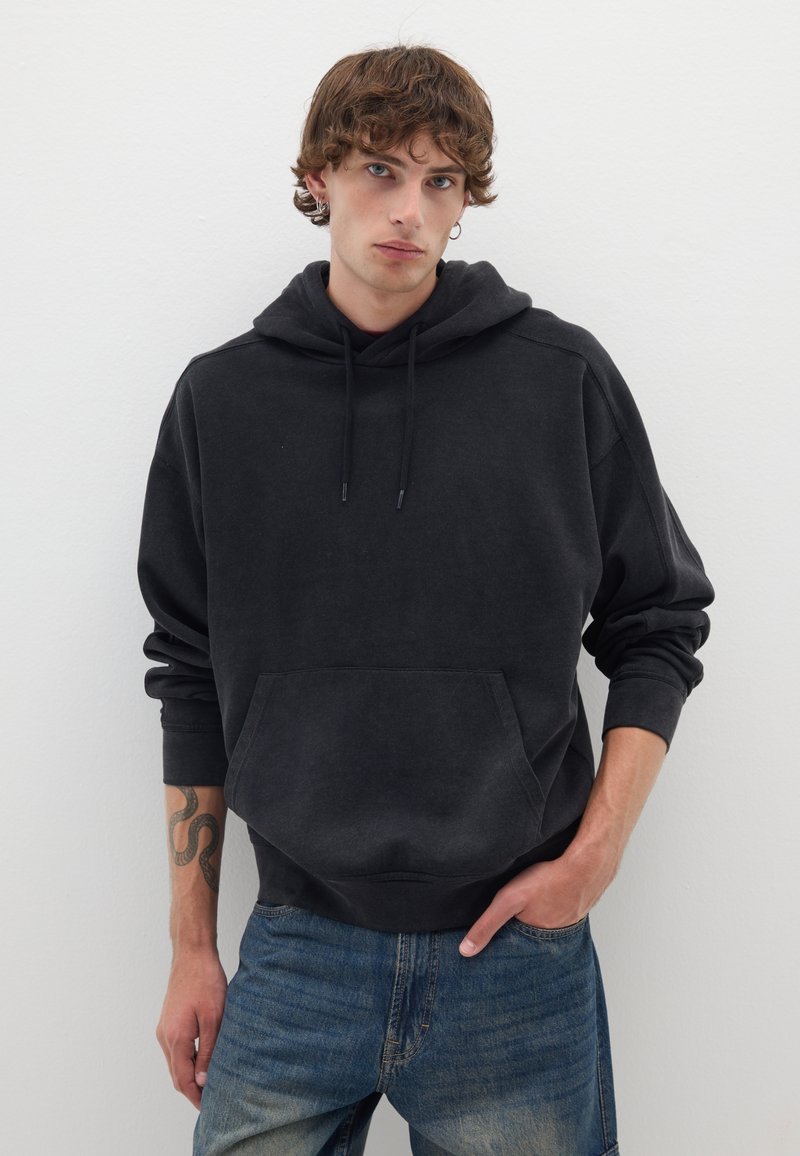 Felpa nera con tasca a marsupio, cappuccio con stringa e polsini a costine. Indossata sopra jeans blu sbiaditi con una vestibilità casual. Tessuto texturizzato.