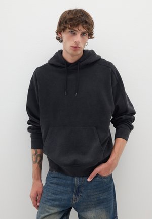 Sudadera negra con bolsillo tipo canguro, capucha con cordón y puños acanalados. Llevada sobre unos vaqueros azules desgastados con un ajuste casual. Tejido texturizado.
