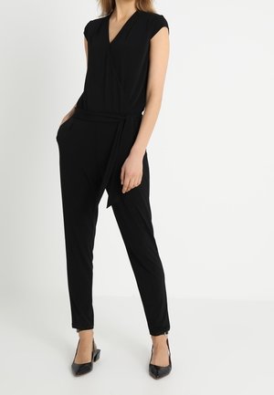Kvinde iført en sort ærmeløs wrap jumpsuit med bælte og sorte spidse hæle, stående mod en helt hvid baggrund.