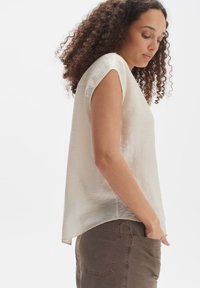 Cremfarbener Kurzarmoberteil mit lockerer Passform, weicher Textur, abgerundetem Saum, kombiniert mit braunen Hosen mit einer Gesäßtasche.