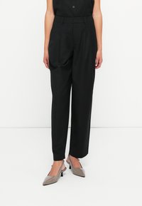 Pantaloni sartoriali neri a vita alta, con pinces, tasche laterali e una texture liscia. Abbinati a tacchi a scomparsa grigi.