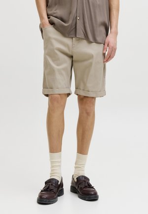 Persona con pantalones cortos beige hasta la rodilla, camisa marrón holgada, calcetines color crema y mocasines marrón oscuro con borlas, de pie con una mano en el bolsillo.