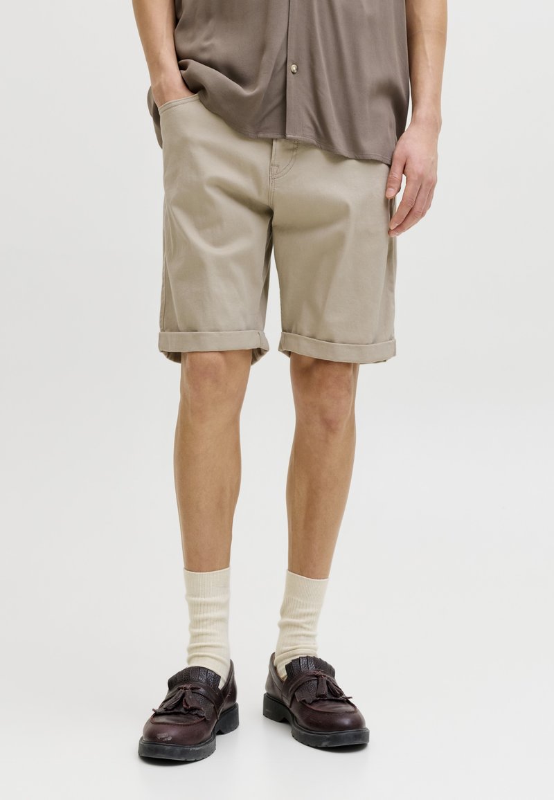 Personne portant un short beige jusqu'aux genoux, une chemise marron ample, des chaussettes couleur crème et des mocassins à pampilles marron foncé, debout avec une main dans la poche.