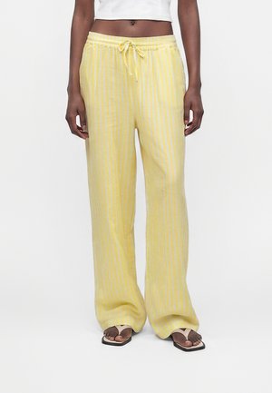 Person iført løse gule bukser med hvide lodrette striber, hvid kort top og beige flipflops, stående på en ensfarvet hvid baggrund.