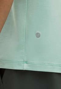 Mėtų žalia sportinė marškinėliai su lygiu paviršiumi, išlenkta apatine dalimi ir Lululemon logotipu pilkoje spalvoje apatiniame kairiajame kampe.