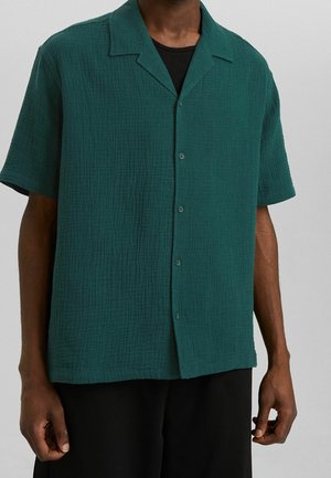 Hombre con una camisa texturizada de manga corta y botones de color verde oscuro sobre una camisa negra, de pie frente a un fondo liso.