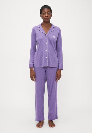 Kvinna stående barfota i en lila långärmad pyjamatröja med knappar och matchande byxor med vit kant och en logotyp på bröstfickan.