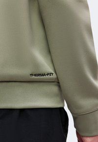 Gros plan d'une personne portant une veste Therma-Fit vert clair avec un pantalon noir, montrant le logo Therma-Fit brodé sur l'ourlet de la veste.