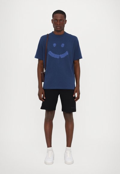 T-shirt en coton bleu marine avec un design graphique de smiley, associé à un short noir et des baskets blanches. Coupe simple et décontractée.