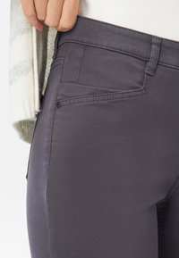 Grå denimbukser med en glat tekstur, der har en sidelomme og en subtil skinnende finish. Taljen inkluderer bæltestropper og syningsdetaljer.