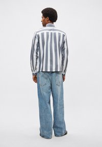 Ριγέ βαμβακερό πουκάμισο με κάθετες γκρι και λευκές γραμμές, συνδυασμένο με φαρδιά ανοιχτόχρωμα τζιν παντελόνια· εμφανείς πίσω τσέπες και ξεθωριασμένη υφή.