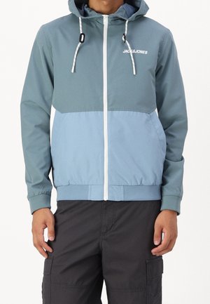 Homme portant un sweat à capuche zippé bicolore bleu avec le logo "JACK & JONES" sur la poitrine et un pantalon cargo noir, debout devant un fond uni.