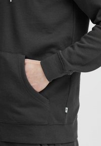 Hoodie noir en mélange de coton doux, doté d'une poche kangourou et de poignets côtelés. Design simpliste avec une étiquette de marque minimaliste sur le côté.