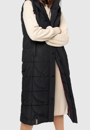 Personne portant un long gilet matelassé noir sans manches sur une robe beige à manches longues, les bras croisés devant un fond uni.