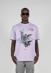 Lavendel kortærmet t-shirt med et grafisk print af en engel og en figur, med tekst og stregkodeoplysninger i en kontrasterende mørk farve.