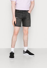 Un joven se encuentra de pie frente a un fondo liso, vistiendo una camiseta lila, pantalones cortos de mezclilla negros con detalles desgastados y zapatillas negras con suelas blancas.
