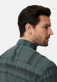 Grünes kariertes Hemd mit Kragen, das horizontale und vertikale Streifen aufweist und aus gewebtem Stoff besteht. Mit einer Schlaufe auf der Rückseite.