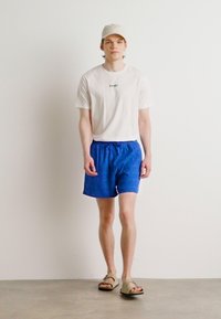 Camiseta blanca con el texto "GANT", pantalones cortos azules y sandalias beige. Modelo que lleva una gorra de color claro posando frente a un fondo liso.
