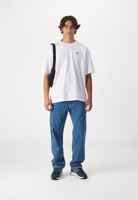 Hvid bomulds t-shirt med sort logo, afslappede blå denim jeans og sorte/grønne sneakers. Model holder en sort skuldertaske.