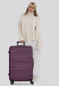 Mujer rubia con un suéter de cuello alto color crema y pantalones blancos, de pie junto a una gran maleta rígida de textura púrpura, con ruedas y asa retráctil.