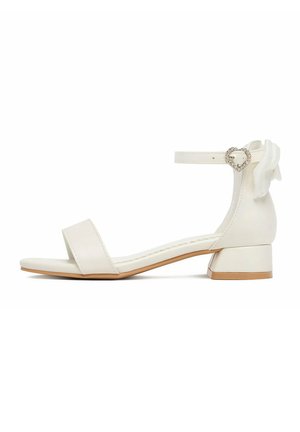 Biała sandałka z odkrytymi palcami, niskim obcasem na szerokim klocku, paskiem wokół kostki ozdobionym klamrą w kształcie serca z kryształków oraz dekoracyjną kokardą z tyłu.