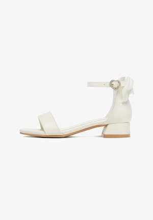 Bílá sandála s otevřenou špičkou, nízkým blokovým podpatkem, kotníkovým páskem s přezkou ve tvaru srdce s kamínky a dekorativní mašlí vzadu.