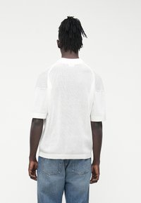 Biała koszula z krótkim rękawem o fakturowanym, żebrowanym wzorze. Zaokrąglony dekolt, dziany wzór i regularny krój. Noszona z niebieskimi dżinsami.