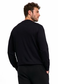 Camicia nera a maniche lunghe realizzata in tessuto morbido, con scollatura rotonda e vestibilità rilassata. Il modello è mostrato di spalle con pantaloni.