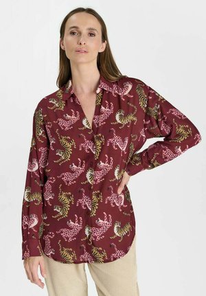 Frau trägt ein burgunderfarbenes Langarmshirt mit pinken und gelben Tiermustern, kombiniert mit hellbeigen Hosen, steht mit einer Hand an der Hüfte.